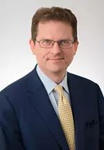 Professional headshot of a man wearing glasses, a navy blue suit jacket, a light blue dress shirt, and a patterned yellow tie. He is facing the camera with a slight smile against a light gray studio background.