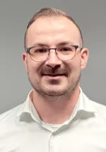 Professional headshot of a man with glasses, short hair, and light facial hair, facing the camera. He is wearing a white collared shirt and is photographed against a neutral gray background, creating a clean and professional appearance.