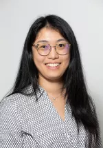 Professional headshot of a woman wearing black glasses and a coral blouse, smiling at the camera against a light background.