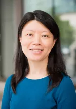 Professional headshot of a woman with shoulder-length dark hair wearing a blue top. She is smiling at the camera in an outdoor setting with a softly blurred background, creating a warm and approachable portrait.