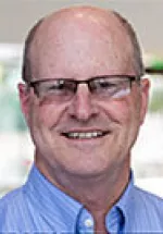 Middle-aged bald man with glasses, smiling, wearing a light blue collared shirt against a blurred green background.