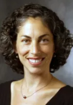 Headshot of a woman with short curly dark hair smiling, wearing a dark top against a neutral studio background.