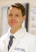 Headshot of a man in a white medical coat and tie, smiling slightly in a clinical hallway setting.