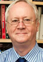 Older man with glasses, wearing a striped shirt and tie, seated in front of a bookshelf background.