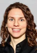 Headshot of a woman with shoulder-length curly hair smiling, wearing a dark top against a neutral background.