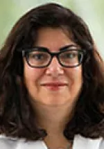 Woman with dark shoulder-length hair and glasses, wearing a white coat, looking directly at the camera with a neutral expression.