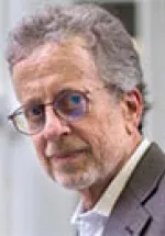 Older man with gray hair and beard, wearing glasses and a blazer, looking slightly to the side against a soft indoor background.