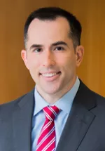 Professional headshot of a smiling man wearing a dark suit jacket, light blue dress shirt, and red striped tie. He is facing the camera against a warm-toned background, creating a polished portrait suitable for a leadership or physician profile.