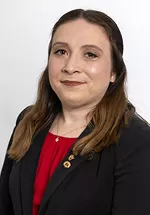 Professional portrait of a woman wearing a black blazer over a red blouse. She is standing against a light gray background and looking at the camera with a slight smile. Small lapel pins are visible on her blazer, giving the image a polished and professional appearance.