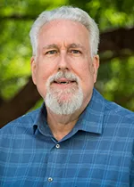 Professional headshot of a man with white hair and a neatly trimmed white beard, wearing a blue collared shirt, standing outdoors with greenery softly blurred in the background.