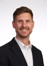 Professional headshot of Adrian Marth, a man with short brown hair and a trimmed beard wearing a dark blazer and white shirt against a light gray background.