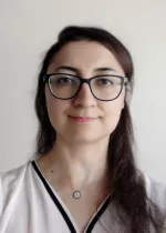Headshot of Zehra Akkaya, a woman with dark hair wearing black glasses and a white blouse against a light background.
