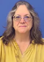 Portrait of a middle-aged woman with shoulder-length wavy gray hair, wearing glasses and a mustard yellow blouse, looking directly at the camera against a plain blue background.