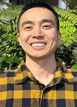 Headshot of a smiling man with short dark hair, standing outdoors in bright sunlight. He is wearing a yellow and black plaid button-up shirt. Green foliage fills the background, creating a natural setting with soft shadows and vibrant colors.