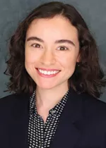 Professional headshot of a smiling person with short curly dark hair wearing a navy blazer and patterned blouse against a gray-blue background.