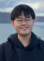 Portrait of a man wearing glasses and a dark hoodie, smiling slightly while standing outdoors. A body of water and cloudy sky are visible in the softly blurred background.