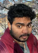 Headshot of a man with dark curly hair and short facial hair, looking toward the camera with a slight smile. He is wearing a maroon hoodie over a gray shirt and a necklace. The photo was taken outdoors, with flowering tree branches and a softly blurred background behind him.