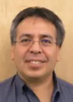 Head-and-shoulders portrait of a man wearing glasses and a dark collared shirt, smiling at the camera. The photo is taken against a light wood-paneled background.