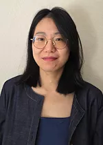 Portrait of a person with shoulder length dark hair and round glasses, wearing a navy blue jacket over a blue top. They are smiling slightly while facing the camera against a plain light colored wall.
