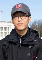Portrait of a person wearing black framed glasses, a gray baseball cap with a red and white "O" logo, and a black jacket with backpack straps visible. They are facing the camera with a neutral expression outdoors, with the White House and an American flag visible in the background.
