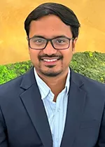 Professional headshot of a person with short dark hair, glasses, and a trimmed beard and mustache, smiling at the camera. They are wearing a navy blazer over a light blue collared shirt. The background is a warm golden gradient with blurred green foliage.