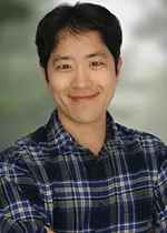 Professional headshot of a smiling person with short black hair, wearing a blue plaid button-down shirt. The background is softly blurred in shades of gray and green, keeping the focus on the subject.