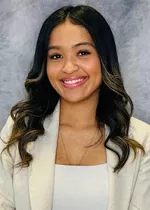 Professional headshot of a smiling person with long, dark wavy hair featuring light highlights, wearing a cream blazer over a white top, against a blue-gray studio background.