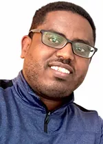 Head-and-shoulders portrait of a person smiling at the camera, wearing rectangular glasses and a navy quarter-zip pullover. The background is plain light gray, with the person's head slightly tilted toward the camera.
