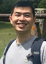 Outdoor head-and-shoulders photo of a person smiling at the camera, wearing a gray T-shirt and a black backpack. A stone railing and a pond with greenery are visible in the background.