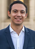 Head-and-shoulders portrait of a person smiling at the camera, wearing a navy blazer over a white collared shirt. The background is softly blurred, showing light-colored stone architecture.