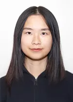Head-and-shoulders portrait of a person with long, straight dark hair, looking directly at the camera with a neutral expression. They are wearing a black zip-up top against a plain light background.