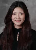 Professional headshot of a person with long, dark brown hair, wearing a black top with a decorative neckline, smiling gently at the camera against a gray studio background.