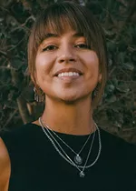 Professional headshot of a smiling person with dark brown hair and bangs, wearing a sleeveless black top, layered silver necklaces, and dangling earrings. The background features softly blurred greenery, creating a warm, natural setting.