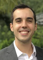 Headshot of Mateus wearing a gray blazer and white shirt, smiling outdoors with greenery in the background.