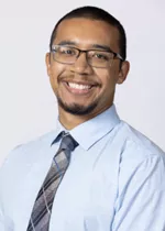 Smiling person wearing glasses, a light blue dress shirt, and a gray plaid tie against a light gray background.