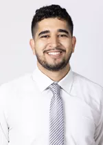 Smiling person with short curly dark hair and trimmed facial hair, wearing a white dress shirt and a striped gray tie against a light gray background.