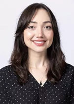 Smiling person with shoulder-length dark brown hair, wearing a black blouse with a subtle dot pattern against a light gray background.