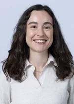 Smiling person with long dark wavy hair, wearing a white collared blouse against a light gray background.