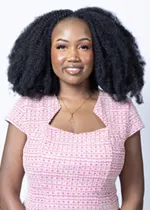Smiling person with natural curly hair, wearing a pink patterned top and a necklace against a light gray background.