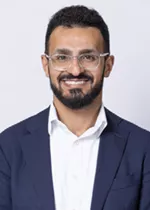 Smiling person with short curly hair, a beard, and glasses, wearing a navy blazer over a white shirt against a light gray background.