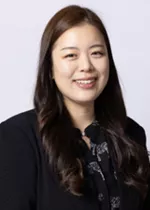Smiling person with long dark hair, wearing a black blazer over a patterned blouse against a light gray background.