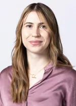 Person with long blonde hair, wearing a mauve satin blouse and a necklace against a light gray background.