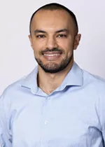 Smiling person with closely cropped hair and trimmed facial hair, wearing a light blue dress shirt against a light gray background.