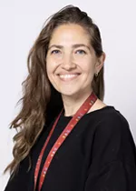 Smiling person with long wavy brown hair, wearing a black top and a red lanyard against a light gray background.
