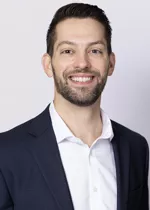 Smiling person with short dark hair and trimmed facial hair, wearing a navy blazer over a white shirt against a light gray background.