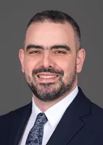 Professional headshot of a smiling man with short dark hair and a neatly trimmed beard, wearing a dark navy suit, white dress shirt, and patterned tie against a plain gray background.