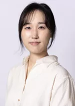 Person with dark hair pulled back, wearing a cream-colored blouse against a light gray background.