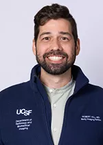 Professional headshot of a smiling person with short dark hair and a beard, wearing a navy UCSF zip-up jacket over a light gray shirt against a light gray background.