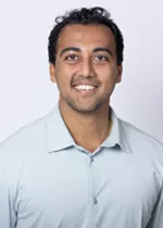 Smiling person with short curly dark hair, wearing a light blue polo shirt against a light gray background.