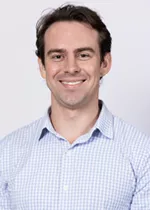 Smiling person with short brown hair, wearing a light blue checkered button-up shirt against a light gray background.